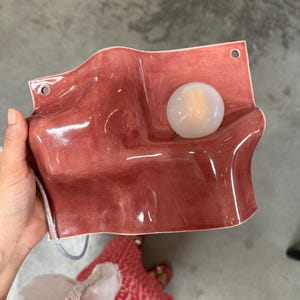 May include: A wall-mounted light fixture featuring a wavy, glossy, dusty rose ceramic base with two circular mounting holes at the top corners and a single, round, frosted white glass globe bulb positioned on the right side of the surface.