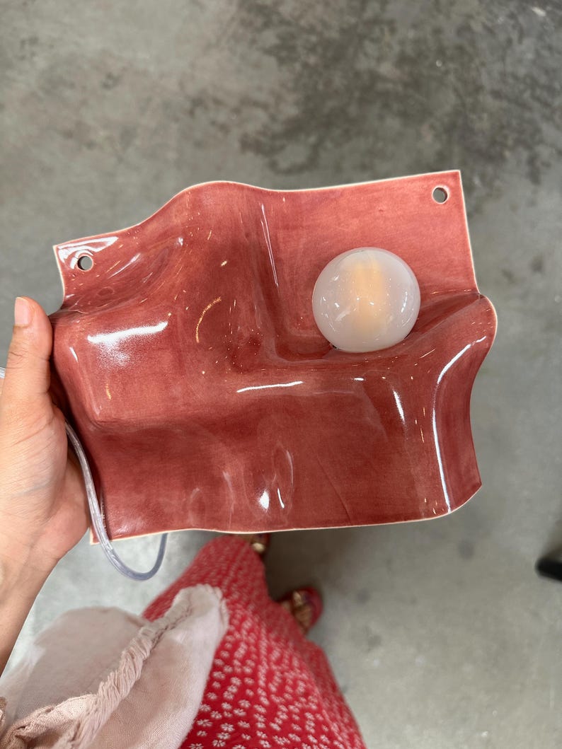 May include: A wall-mounted light fixture featuring a wavy, glossy, dusty rose ceramic base with two circular mounting holes at the top corners and a single, round, frosted white glass globe bulb positioned on the right side of the surface.