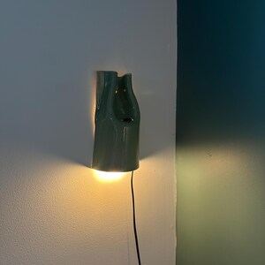 May include: A sage green ceramic wall sconce with a smooth, glossy finish and an organic, asymmetrical shape. The fixture emits a warm, downward-facing light and includes a thin black power cord extending from the bottom of the lamp housing.