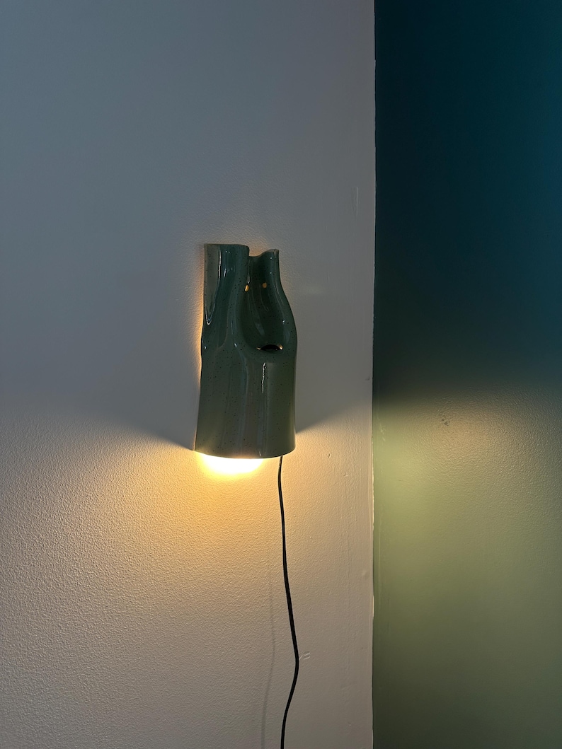 May include: A sage green ceramic wall sconce with a smooth, glossy finish and an organic, asymmetrical shape. The fixture emits a warm, downward-facing light and includes a thin black power cord extending from the bottom of the lamp housing.