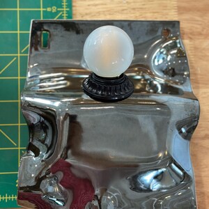 May include: A wall-mounted light fixture featuring a reflective, wavy chrome-finished metal backplate and a single spherical white glass bulb. The fixture is positioned next to a green cutting mat with markings in inches, showing a width of 6 inches.