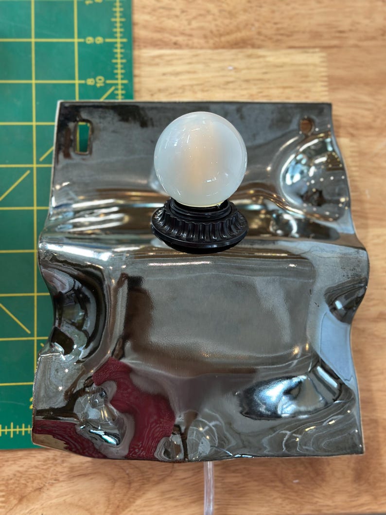 May include: A wall-mounted light fixture featuring a reflective, wavy chrome-finished metal backplate and a single spherical white glass bulb. The fixture is positioned next to a green cutting mat with markings in inches, showing a width of 6 inches.