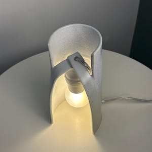 K&ouml;nnte beinhalten: Eine wei&szlig;e Tischleuchte aus Keramik mit einer geschwungenen, zylindrischen Struktur, die eine zentrale Gl&uuml;hbirne umschlie&szlig;t. Die Lampe hat eine glatte, matte Oberfl&auml;che und ein einzelnes Stromkabel, das warmes Licht ausstrahlt.