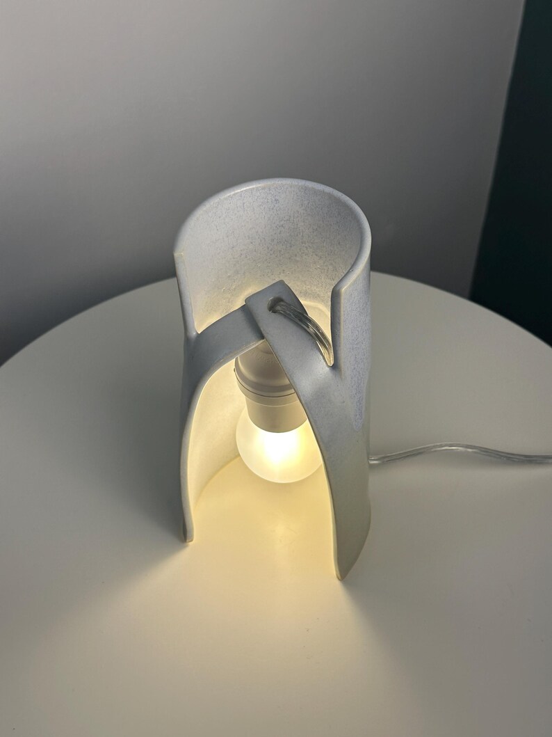 K&ouml;nnte beinhalten: Eine wei&szlig;e Tischleuchte aus Keramik mit einer geschwungenen, zylindrischen Struktur, die eine zentrale Gl&uuml;hbirne umschlie&szlig;t. Die Lampe hat eine glatte, matte Oberfl&auml;che und ein einzelnes Stromkabel, das warmes Licht ausstrahlt.