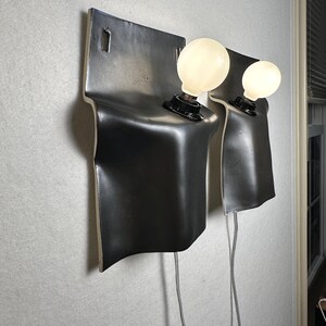 May include: Two wall-mounted sconces featuring a matte black, curved ceramic backplate with a smooth, flowing silhouette. Each fixture includes a single black socket holding a round, glowing white light bulb, with a thin gray power cord extending downward.