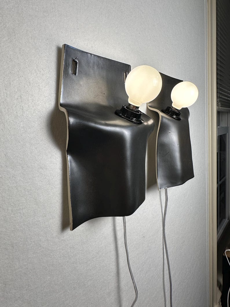 May include: Two wall-mounted sconces featuring a matte black, curved ceramic backplate with a smooth, flowing silhouette. Each fixture includes a single black socket holding a round, glowing white light bulb, with a thin gray power cord extending downward.