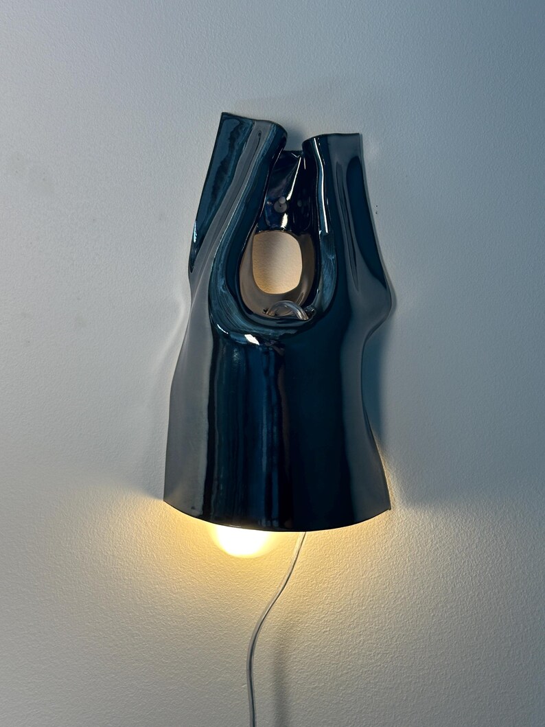 May include: A dark blue, glossy ceramic wall sconce with an organic, fluid shape. The fixture features a central cutout and a downward-facing light bulb that emits a warm glow, with a thin white power cord extending from the bottom of the unit.