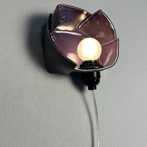 K&ouml;nnte beinhalten: Eine Wandleuchte mit einem dunklen, bl&uuml;tenblattf&ouml;rmigen Keramikschirm und einer gl&auml;nzenden, irisierenden violetten Innenseite. Eine schwarze Fassung h&auml;lt eine kleine, runde, mattwei&szlig;e Gl&uuml;hbirne, die warmes Licht abgibt, verbunden mit einem transparenten Kabel.