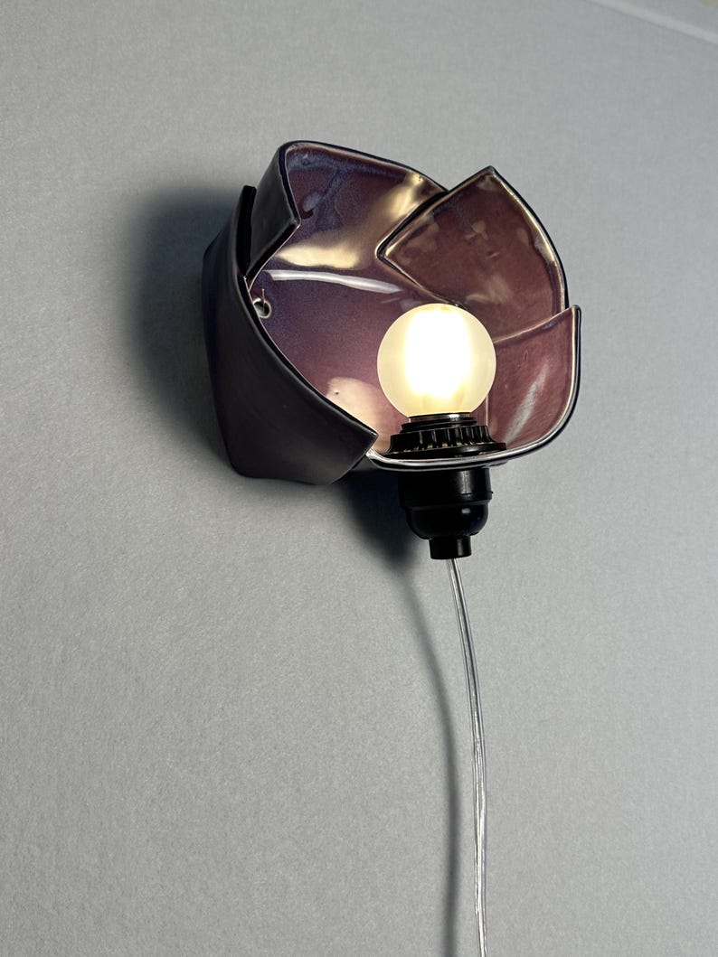 K&ouml;nnte beinhalten: Eine Wandleuchte mit einem dunklen, bl&uuml;tenblattf&ouml;rmigen Keramikschirm und einer gl&auml;nzenden, irisierenden violetten Innenseite. Eine schwarze Fassung h&auml;lt eine kleine, runde, mattwei&szlig;e Gl&uuml;hbirne, die warmes Licht abgibt, verbunden mit einem transparenten Kabel.