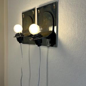 May include: Two wall-mounted sconces with a dark metallic finish. Each sconce features a round, exposed light bulb and a curved shelf. The sconces are mounted on a light-colored wall, with the electrical cords visible.