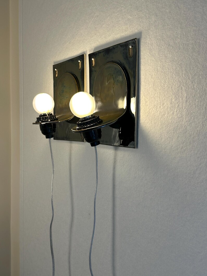 May include: Two wall-mounted sconces with a dark metallic finish. Each sconce features a round, exposed light bulb and a curved shelf. The sconces are mounted on a light-colored wall, with the electrical cords visible.