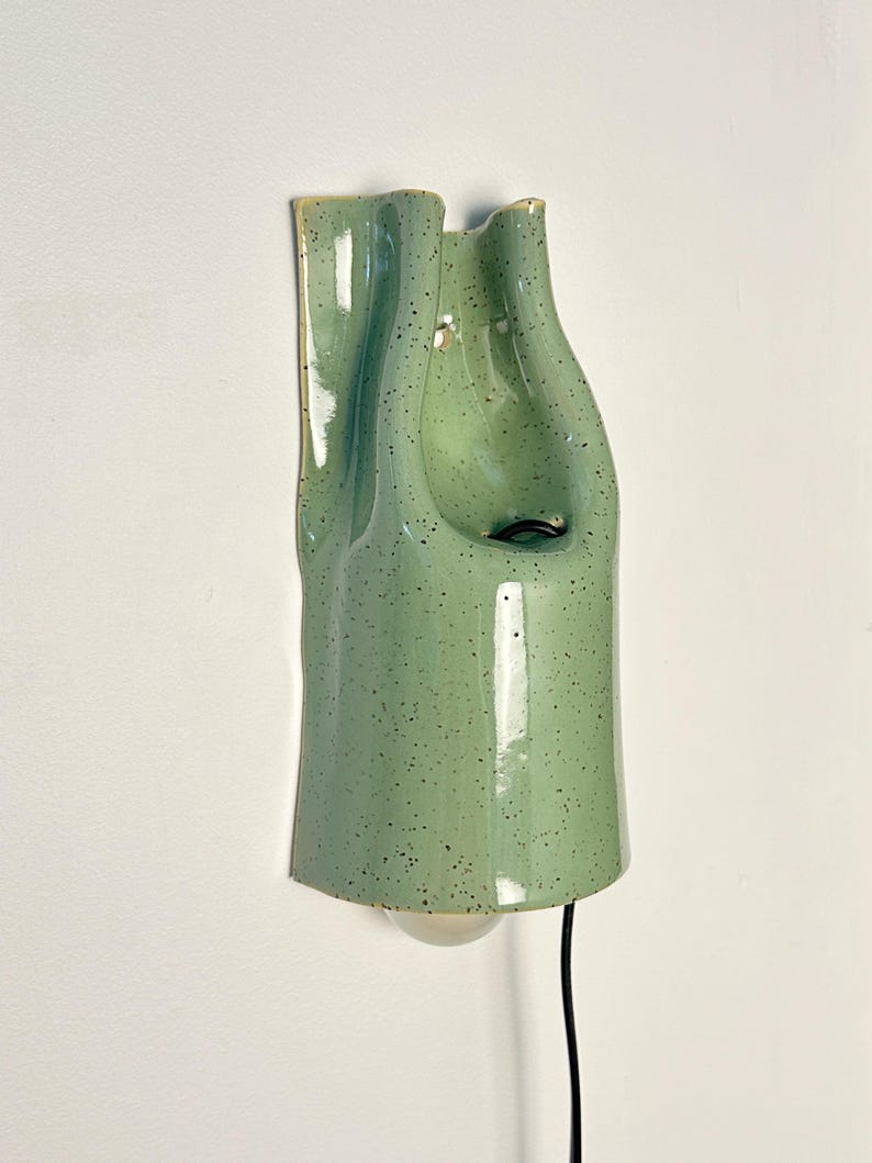 May include: A wall-mounted ceramic light fixture with a glossy, light green glaze and dark speckles. The piece features an organic, folded, and undulating shape with a central opening for a light bulb and a black electrical cord extending downward.