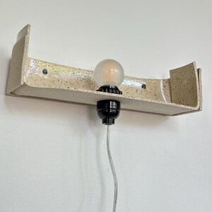 May include: A speckled, cream-colored ceramic wall sconce with a curved shelf design. It features a black light socket and a frosted bulb. The sconce is mounted on a white wall, with a clear electrical cord.