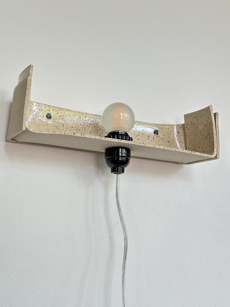 May include: A speckled, cream-colored ceramic wall sconce with a curved shelf design. It features a black light socket and a frosted bulb. The sconce is mounted on a white wall, with a clear electrical cord.