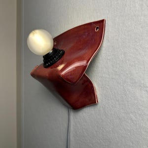 Puede incluir: Un aplique de pared con una base de cer&aacute;mica de color rojo oscuro brillante y forma irregular plegada. Un casquillo negro sostiene una bombilla redonda de color blanco esmerilado. Un cable el&eacute;ctrico fino y claro cuelga hacia abajo.