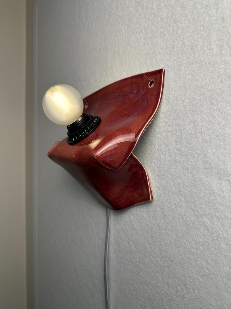 Puede incluir: Un aplique de pared con una base de cer&aacute;mica de color rojo oscuro brillante y forma irregular plegada. Un casquillo negro sostiene una bombilla redonda de color blanco esmerilado. Un cable el&eacute;ctrico fino y claro cuelga hacia abajo.