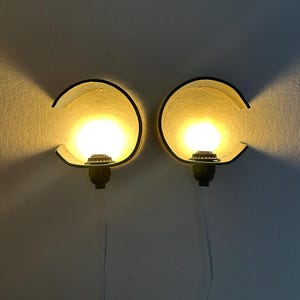 May include: A pair of wall-mounted lamps featuring curved, dark-colored semi-circular shades that partially enclose a glowing, warm-toned spherical light bulb. Each lamp is attached to a black base and includes a thin, transparent power cord.