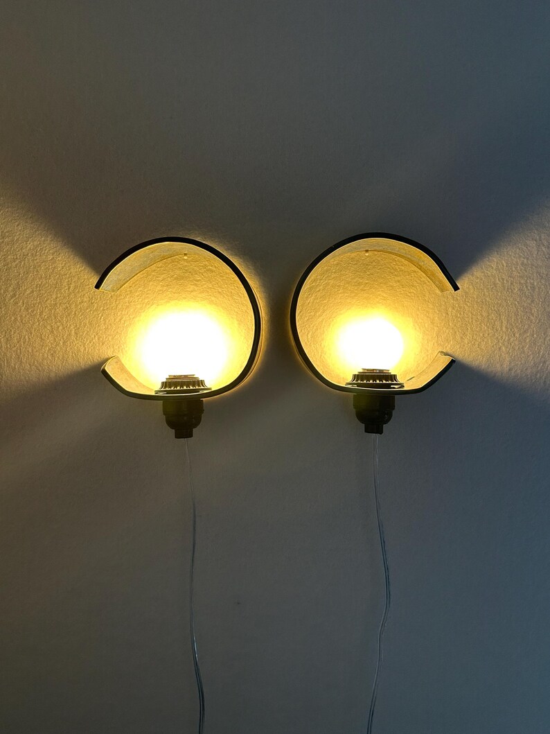 May include: A pair of wall-mounted lamps featuring curved, dark-colored semi-circular shades that partially enclose a glowing, warm-toned spherical light bulb. Each lamp is attached to a black base and includes a thin, transparent power cord.