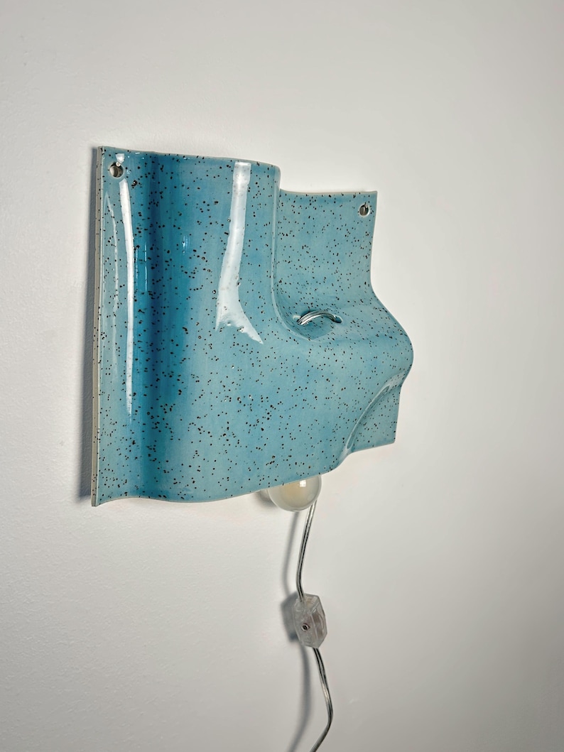 May include: A wall-mounted light fixture made of light blue ceramic with a dark speckled pattern. The fixture has a curved, sculptural form with two small holes for mounting and a single light bulb extending from the bottom with a clear power cord.