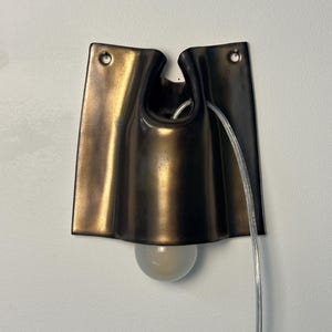 May include: A wall-mounted sconce featuring a dark bronze-toned, curved metal shade with a smooth, folded design. The fixture includes two mounting holes at the top and a clear electrical cord extending from the center, with a round light bulb.