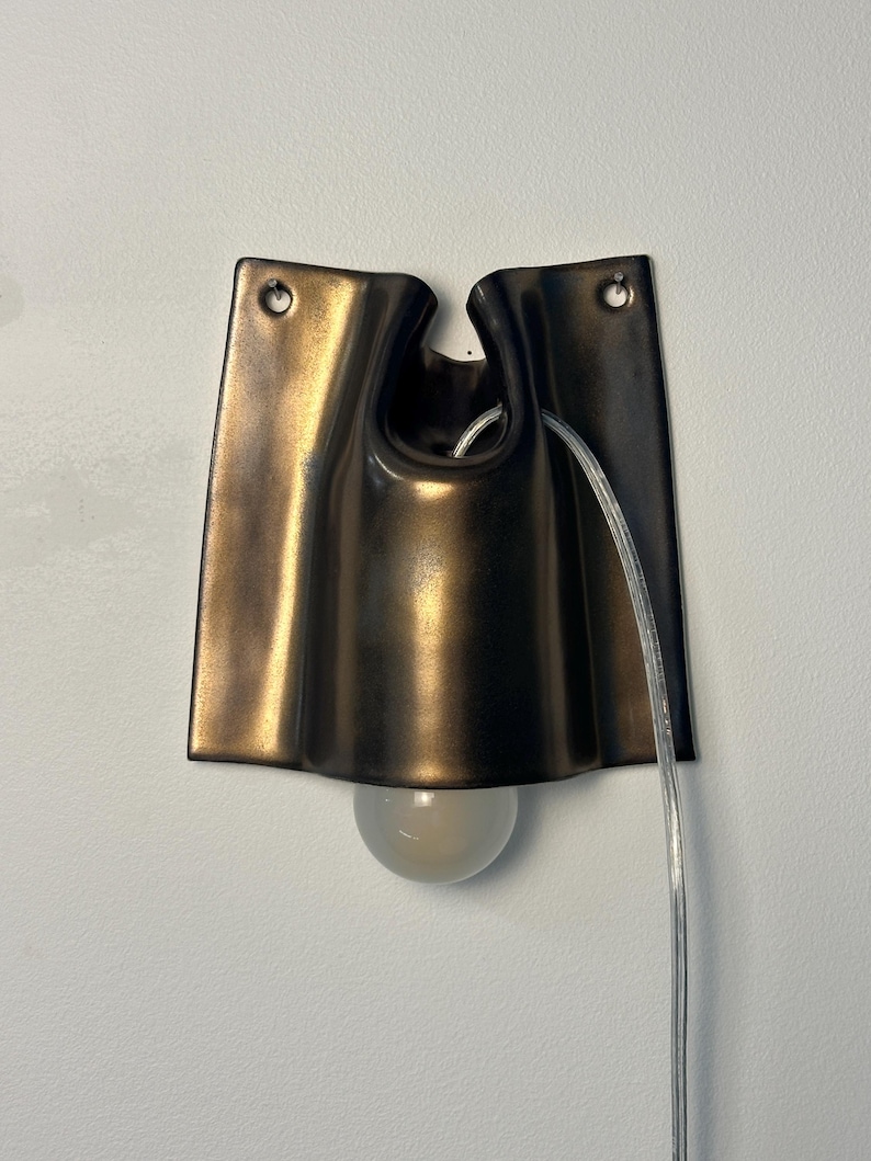 May include: A wall-mounted sconce featuring a dark bronze-toned, curved metal shade with a smooth, folded design. The fixture includes two mounting holes at the top and a clear electrical cord extending from the center, with a round light bulb.