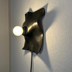 May include: A black wall-mounted lamp featuring a curved, organic-shaped backplate with a smooth finish. A single round, glowing light bulb is attached to the center of the plate. A thin black power cord extends downward from the base of the fixture.