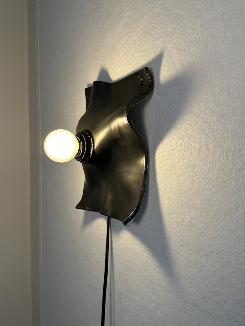 May include: A black wall-mounted lamp featuring a curved, organic-shaped backplate with a smooth finish. A single round, glowing light bulb is attached to the center of the plate. A thin black power cord extends downward from the base of the fixture.