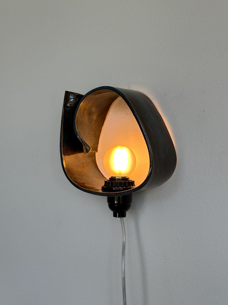May include: A wall-mounted light fixture with a black, curved metal shade and a warm, glowing lightbulb. The interior of the shade is gold-toned, creating a contrast with the dark exterior. A black socket and clear cord complete the design.