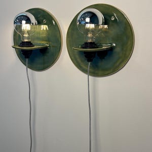 May include: A pair of wall-mounted sconces featuring circular, glossy olive-green ceramic backplates. Each fixture includes a small horizontal ceramic shelf holding a black socket with a clear globe light bulb topped with a reflective silver cap.