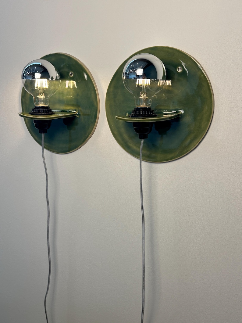 May include: A pair of wall-mounted sconces featuring circular, glossy olive-green ceramic backplates. Each fixture includes a small horizontal ceramic shelf holding a black socket with a clear globe light bulb topped with a reflective silver cap.