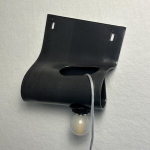 Puede incluir: Una l&aacute;mpara de pared negra con un cuerpo de material flexible y curvado que presenta dos orificios de montaje rectangulares en la parte superior. Una abertura central horizontal permite el paso de un cable transparente hacia una bombilla redonda.