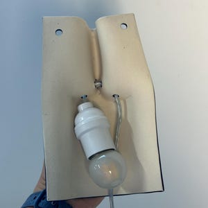 Puede incluir: Un aplique de pared con una placa trasera curva de color blanco roto, hecha de cer&aacute;mica o material mate, con dos orificios de montaje circulares en la parte superior. Un casquillo de pl&aacute;stico blanco est&aacute; fijado en el centro con una bombilla clara.