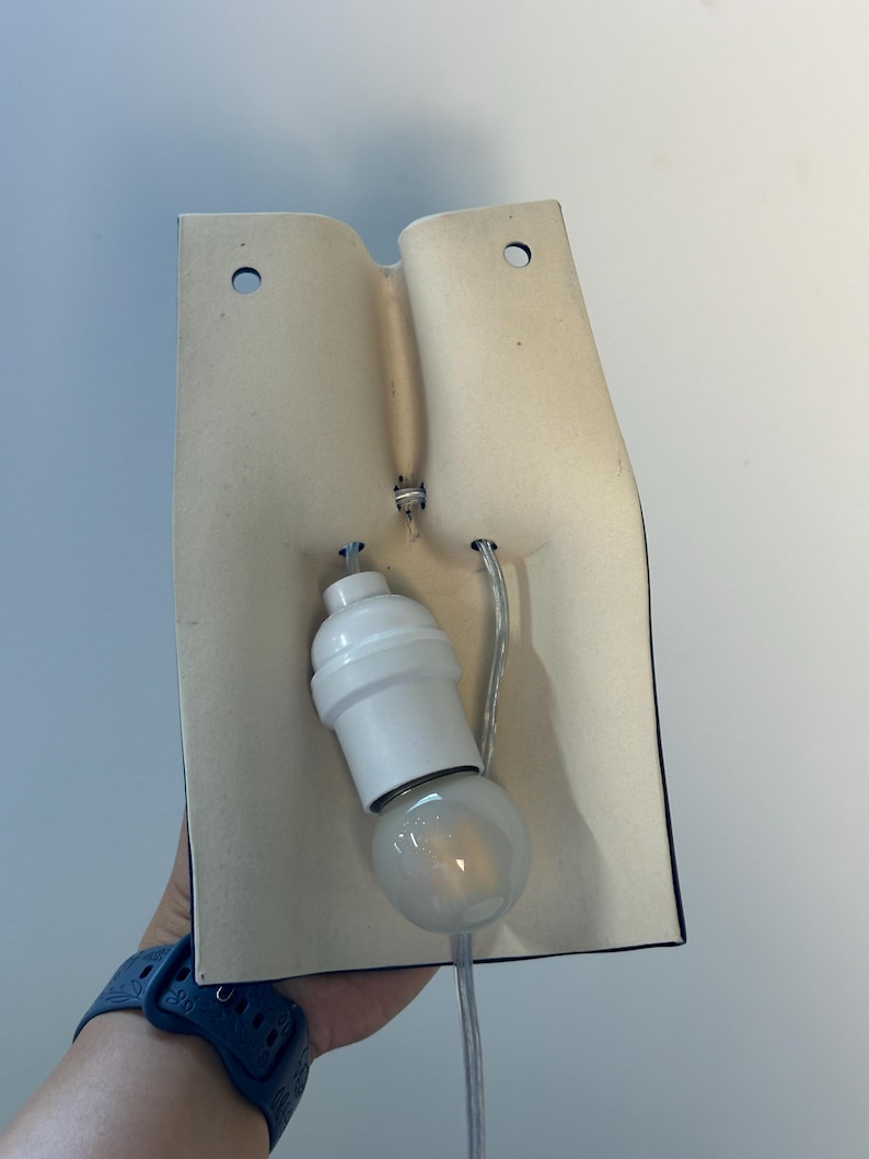 Puede incluir: Un aplique de pared con una placa trasera curva de color blanco roto, hecha de cer&aacute;mica o material mate, con dos orificios de montaje circulares en la parte superior. Un casquillo de pl&aacute;stico blanco est&aacute; fijado en el centro con una bombilla clara.