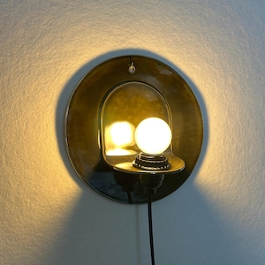 May include: A circular wall sconce featuring a glossy, dark olive-green ceramic backplate with a curved shelf holding a single, glowing spherical light bulb. The fixture includes a black power cord extending downward from the base of the socket.