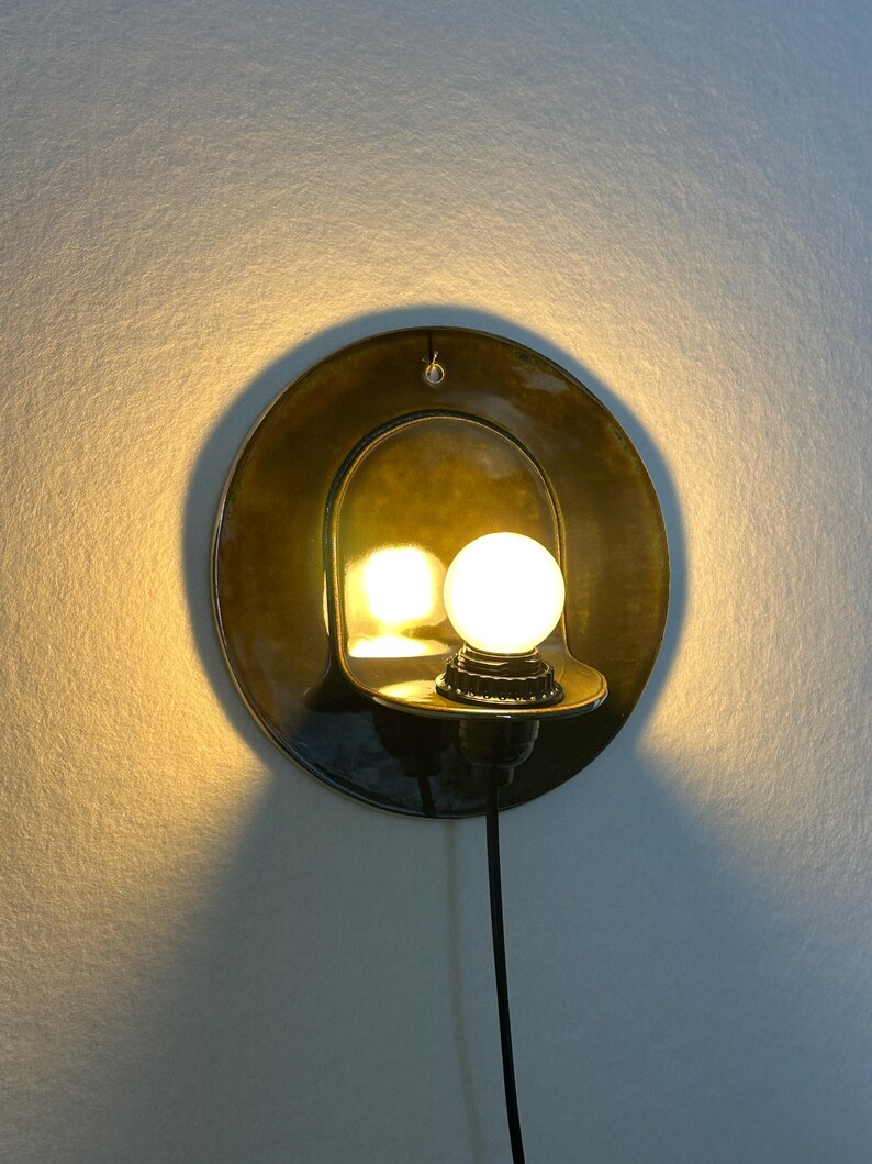 May include: A circular wall sconce featuring a glossy, dark olive-green ceramic backplate with a curved shelf holding a single, glowing spherical light bulb. The fixture includes a black power cord extending downward from the base of the socket.
