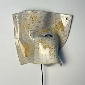 May include: A wall-mounted ceramic light fixture with an irregular, wavy, and folded shape. The surface is off-white with brown speckles and translucent yellow glaze patches. It features two circular mounting holes and a black power cord.