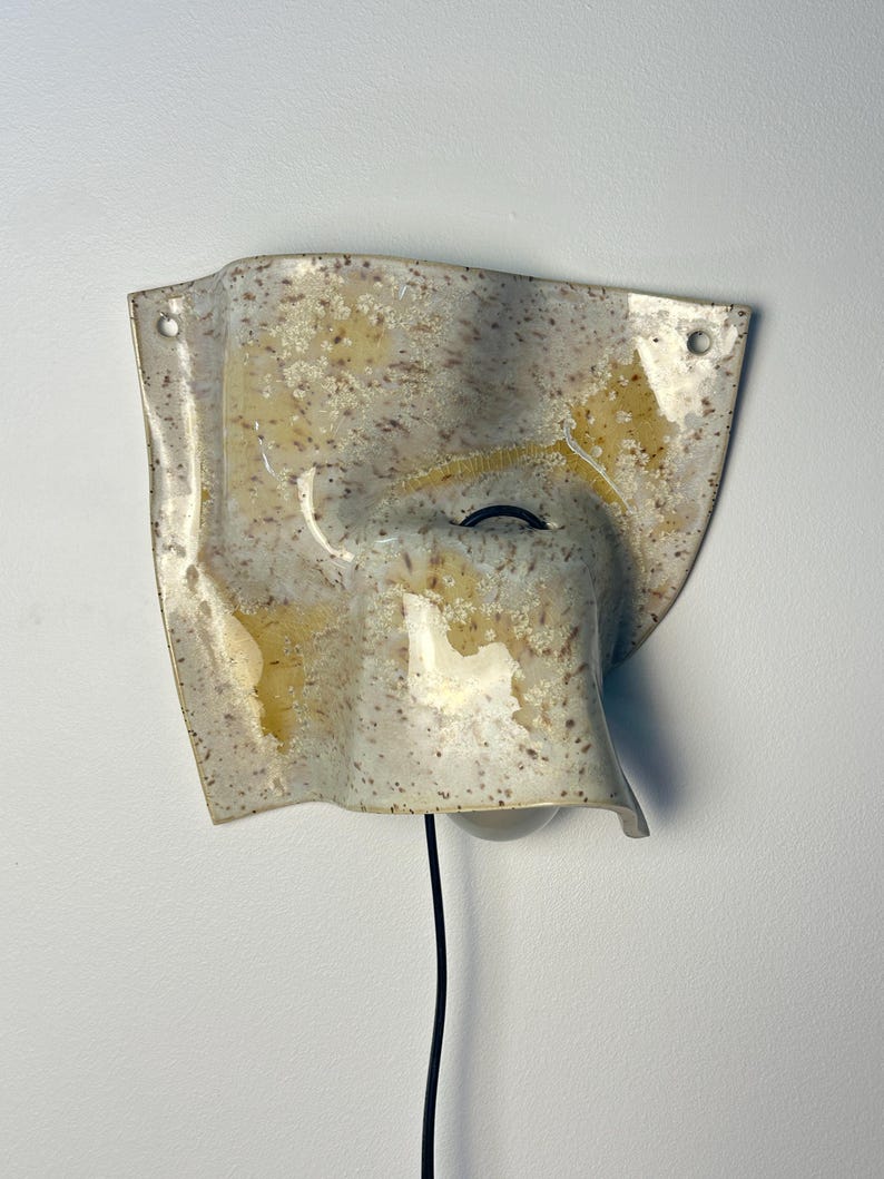 May include: A wall-mounted ceramic light fixture with an irregular, wavy, and folded shape. The surface is off-white with brown speckles and translucent yellow glaze patches. It features two circular mounting holes and a black power cord.