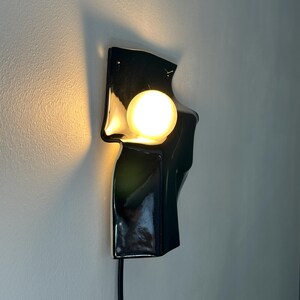 Puede incluir: Un aplique de pared con una base de cer&aacute;mica negra brillante de forma irregular y superficie lisa y reflectante. Una bombilla esf&eacute;rica de luz c&aacute;lida est&aacute; centrada en la forma, proyectando un suave resplandor sobre la pared.
