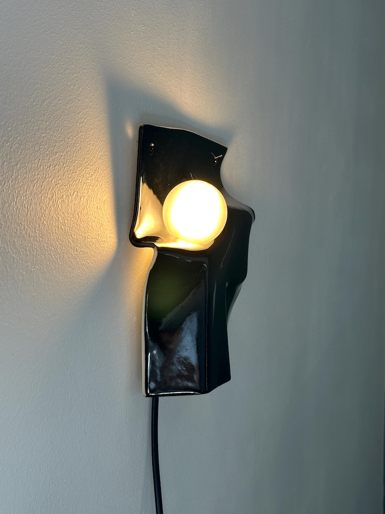 Puede incluir: Un aplique de pared con una base de cer&aacute;mica negra brillante de forma irregular y superficie lisa y reflectante. Una bombilla esf&eacute;rica de luz c&aacute;lida est&aacute; centrada en la forma, proyectando un suave resplandor sobre la pared.