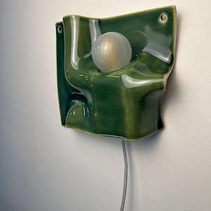 May include: A wall-mounted ceramic light fixture with a glossy, deep green glaze and an undulating, folded surface. A single, spherical, frosted white light bulb is centered within the form. The piece features two small circular holes for mounting.
