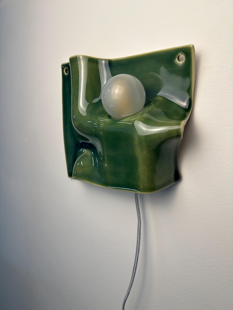 May include: A wall-mounted ceramic light fixture with a glossy, deep green glaze and an undulating, folded surface. A single, spherical, frosted white light bulb is centered within the form. The piece features two small circular holes for mounting.