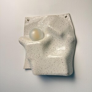 May include: A wall-mounted ceramic light fixture with an off-white, speckled glaze and an irregular, undulating surface. A single, spherical, frosted glass bulb is integrated into the left side of the form, which features two small holes at the top.