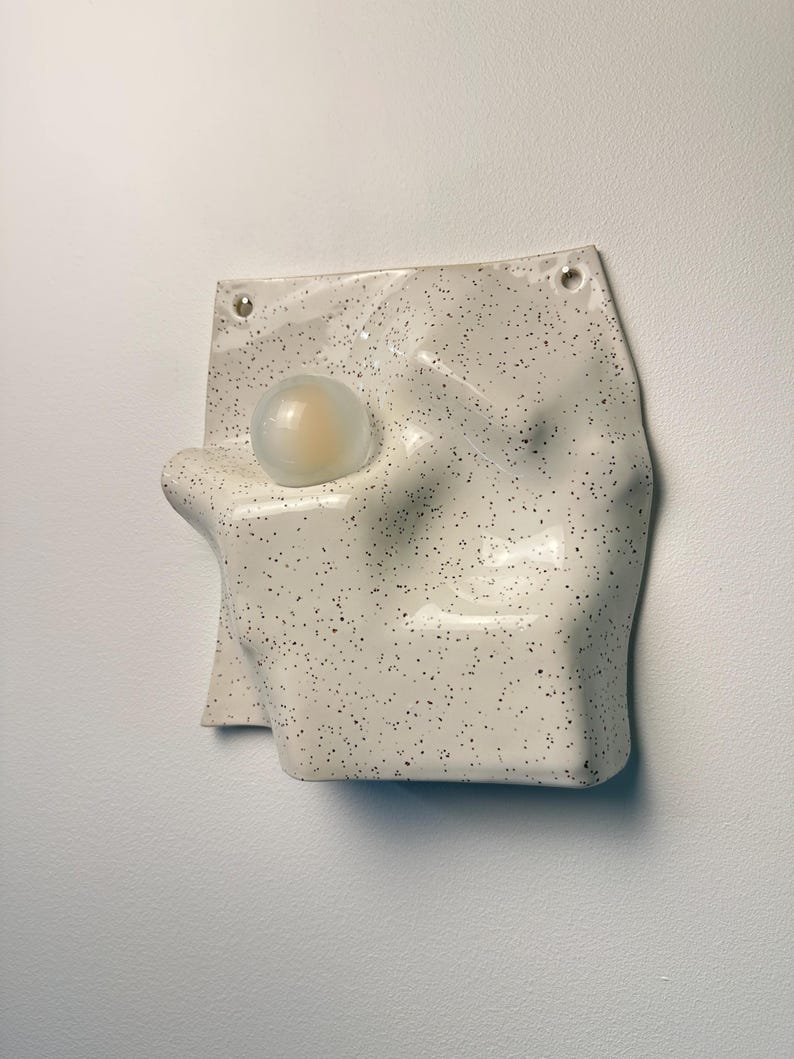 May include: A wall-mounted ceramic light fixture with an off-white, speckled glaze and an irregular, undulating surface. A single, spherical, frosted glass bulb is integrated into the left side of the form, which features two small holes at the top.