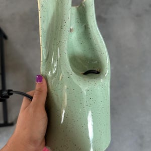 May include: A light green ceramic fountain with a speckled finish and a glossy glaze. The structure features a tall, curved, and organic shape with a central opening and a black electrical cord extending from the base for power connectivity.