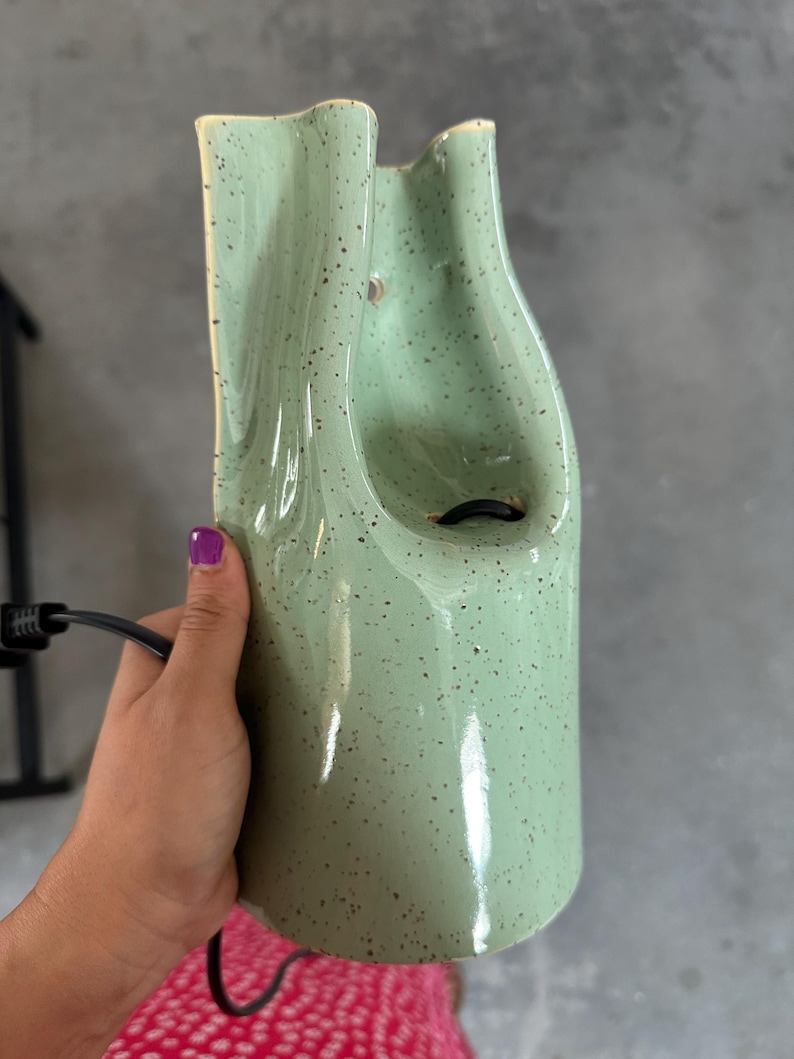 May include: A light green ceramic fountain with a speckled finish and a glossy glaze. The structure features a tall, curved, and organic shape with a central opening and a black electrical cord extending from the base for power connectivity.