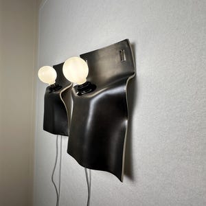 May include: Two wall-mounted lamps featuring curved, matte black rectangular shades with rounded corners. Each lamp has a single round, frosted white light bulb protruding from the center, connected to a light gray power cord hanging downward.