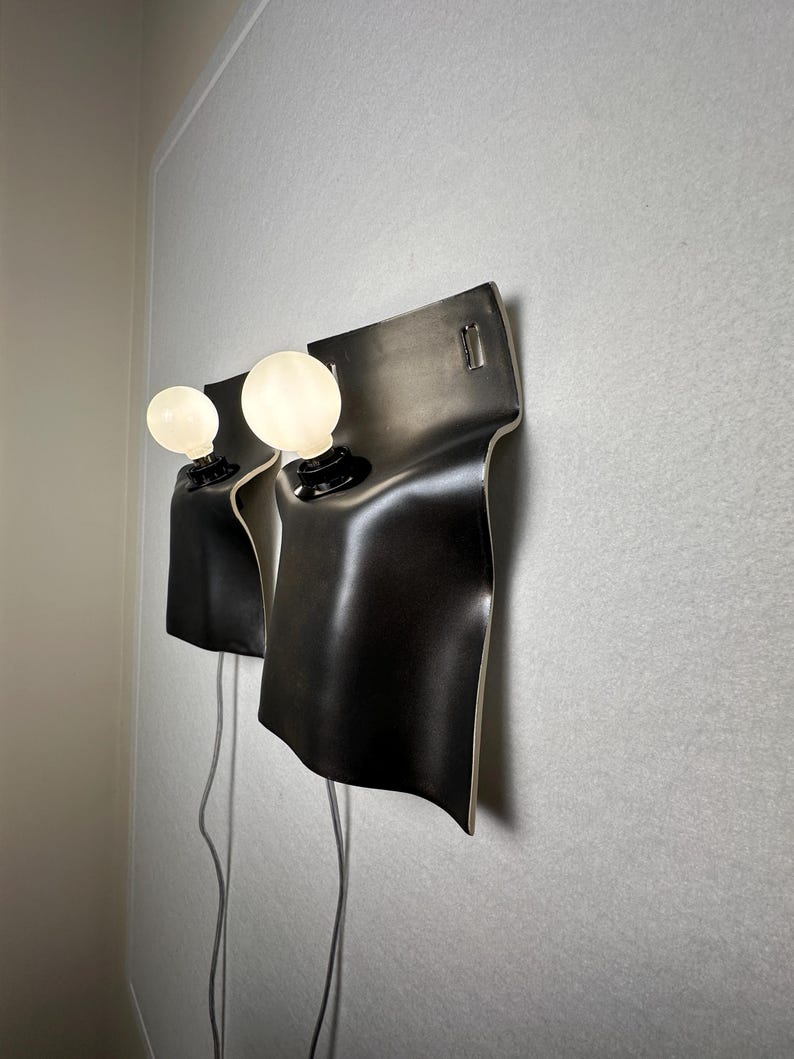 May include: Two wall-mounted lamps featuring curved, matte black rectangular shades with rounded corners. Each lamp has a single round, frosted white light bulb protruding from the center, connected to a light gray power cord hanging downward.