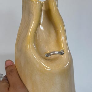 Pu&ograve; includere: Un vaso in ceramica alto, lucido e di colore giallo pallido con una forma irregolare, organica e fluida. L'apertura superiore presenta una profonda rientranza curva e un piccolo tubo di plastica trasparente e flessibile passa attraverso un foro al centro.