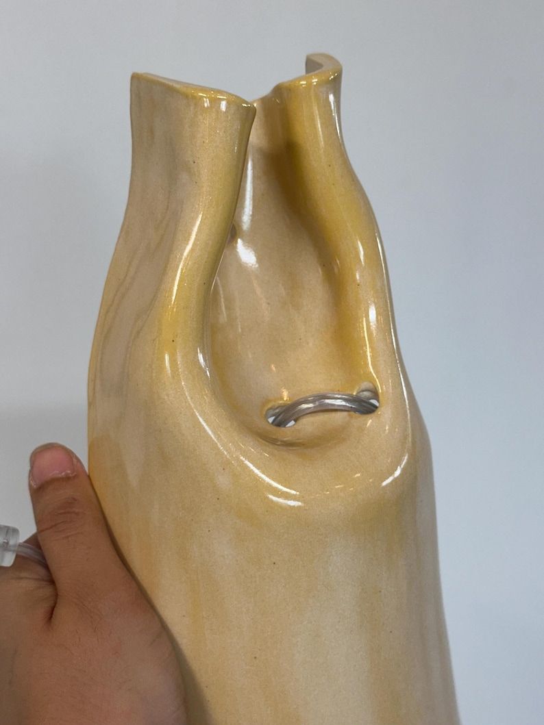 Pu&ograve; includere: Un vaso in ceramica alto, lucido e di colore giallo pallido con una forma irregolare, organica e fluida. L'apertura superiore presenta una profonda rientranza curva e un piccolo tubo di plastica trasparente e flessibile passa attraverso un foro al centro.