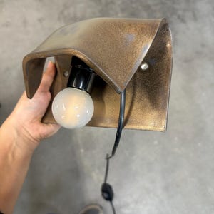 Pode incluir: Um candeeiro de parede com um abajur de metal curvo em tom de bronze com acabamento texturizado. A lumin&aacute;ria inclui um casquilho preto com uma l&acirc;mpada redonda de vidro fosco branco e um cabo de alimenta&ccedil;&atilde;o preto com interruptor.